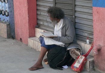 Uomo senza fissa dimora seduto per strada che legge un libro e ha accanto a sé una chitarra rossa